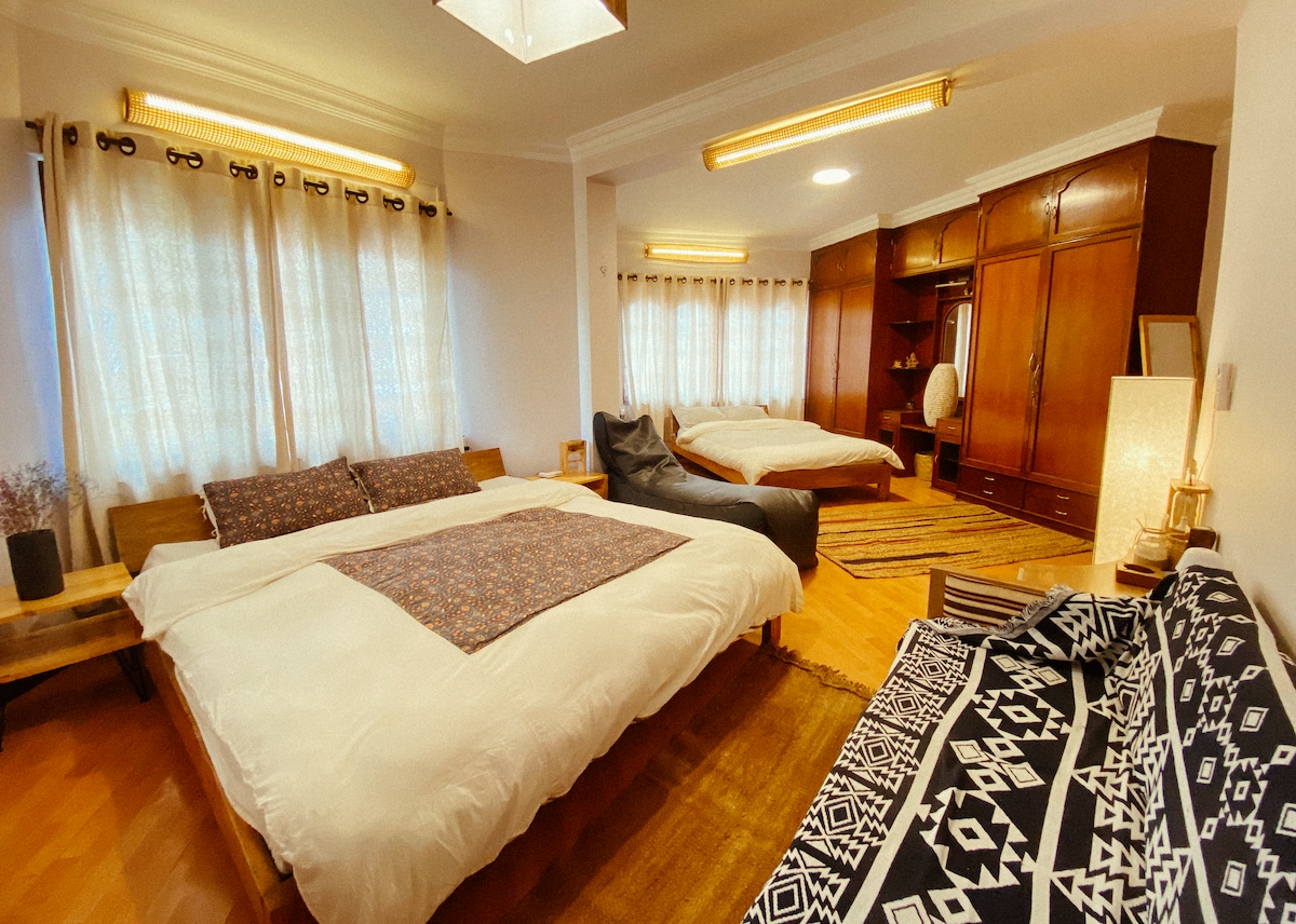 A spacious sleeping area features two large beds dressed in white linens, accented with a patterned throw. Natural light floods the room through multiple windows with sheer curtains. A cozy seating area includes a comfortable chair, and wooden furniture complements the warm wood floors.