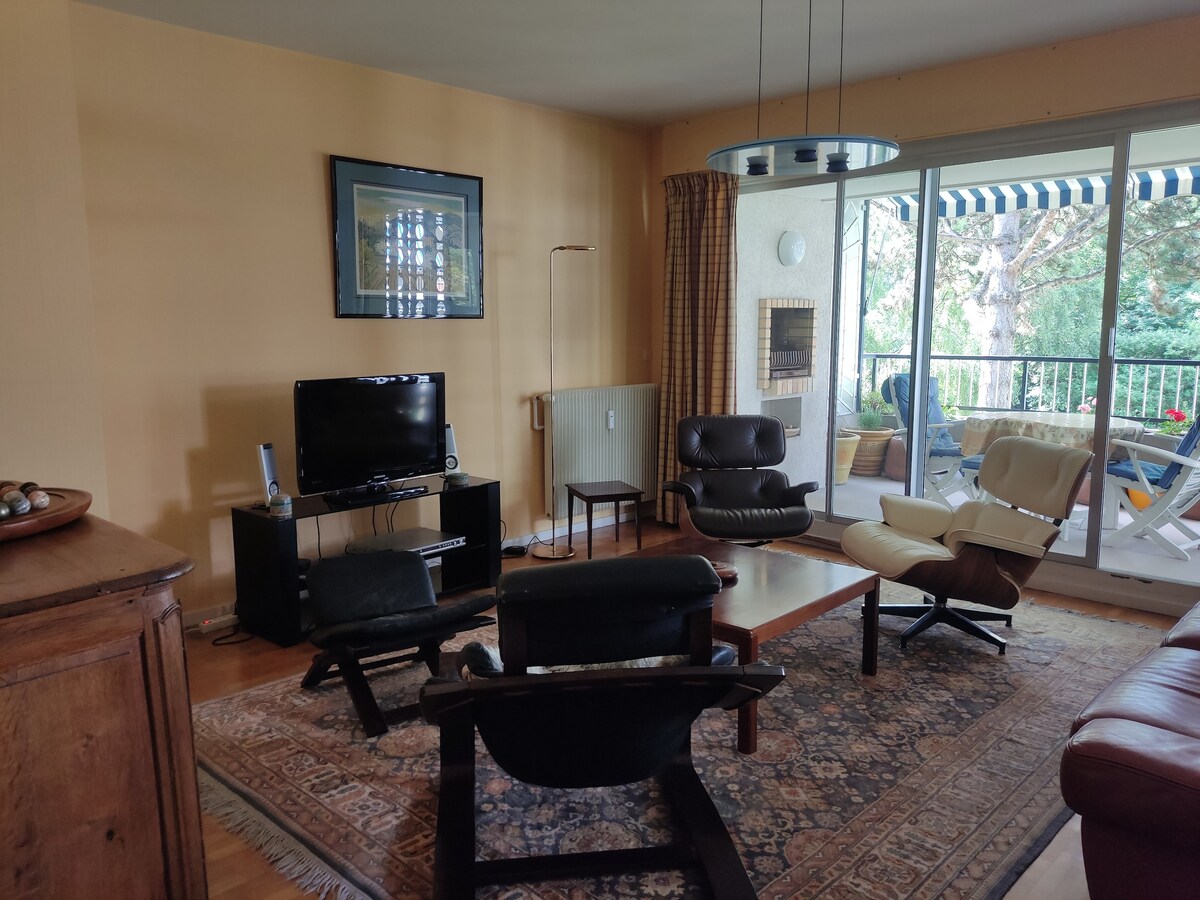 A living area is shown featuring a mix of seating options, including two armchairs and a lounge chair around a central wooden coffee table. A television is positioned on an entertainment stand, and large glass doors open to a balcony view of greenery.