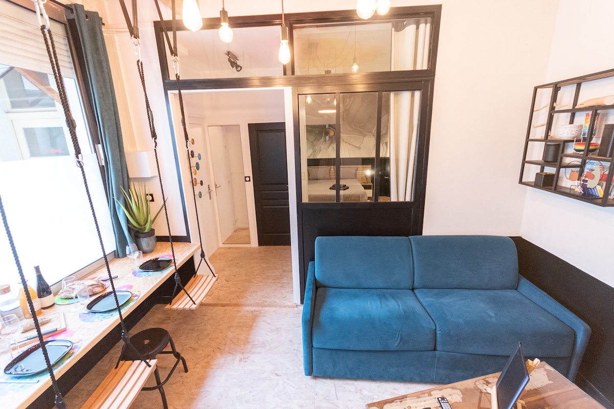 A bright living space is featured, with a cozy blue sofa positioned adjacent to a swinging chair table setup. Natural light is flooding in through large windows, illuminating the artwork on the wall. A dividing glass wall provides a glimpse into the bedroom area.