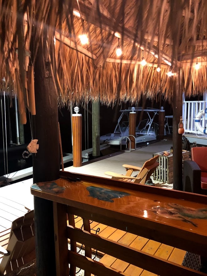A tiki hut with a thatched roof is illuminated by soft lights, providing a cozy space adjacent to a wooden dock. Comfortable seating and a bar area are visible, along with a boat lift and details of the waterway beyond.
