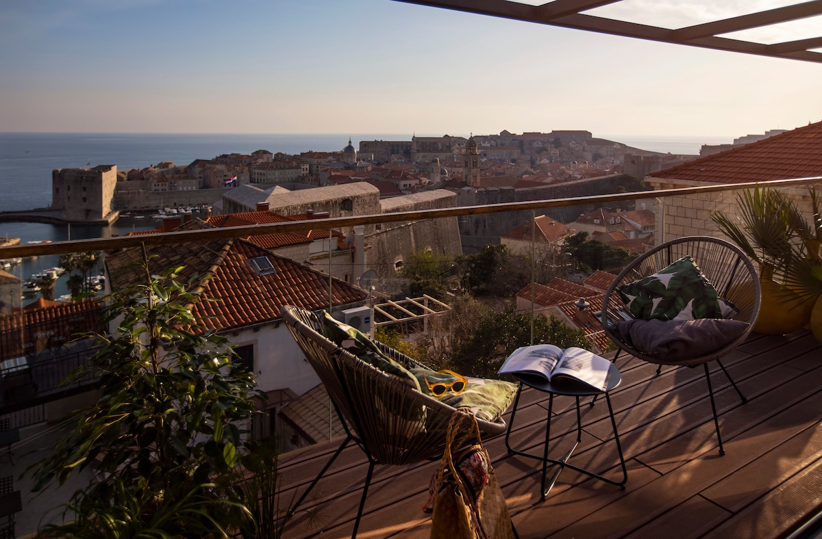 A terrace area features two comfortable chairs positioned towards an expansive view of the Old Town and the sea. Warm sunlight casts gentle shadows on the wooden floor, and a decorative plant adds a touch of greenery, enhancing the outdoor space.