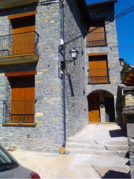 A three-story stone building features wooden balconies and window shutters. The entrance is marked by an arched doorway, leading to a staircase that connects the ground level with the upper floors. Nearby, a car is parked on the gravel driveway.