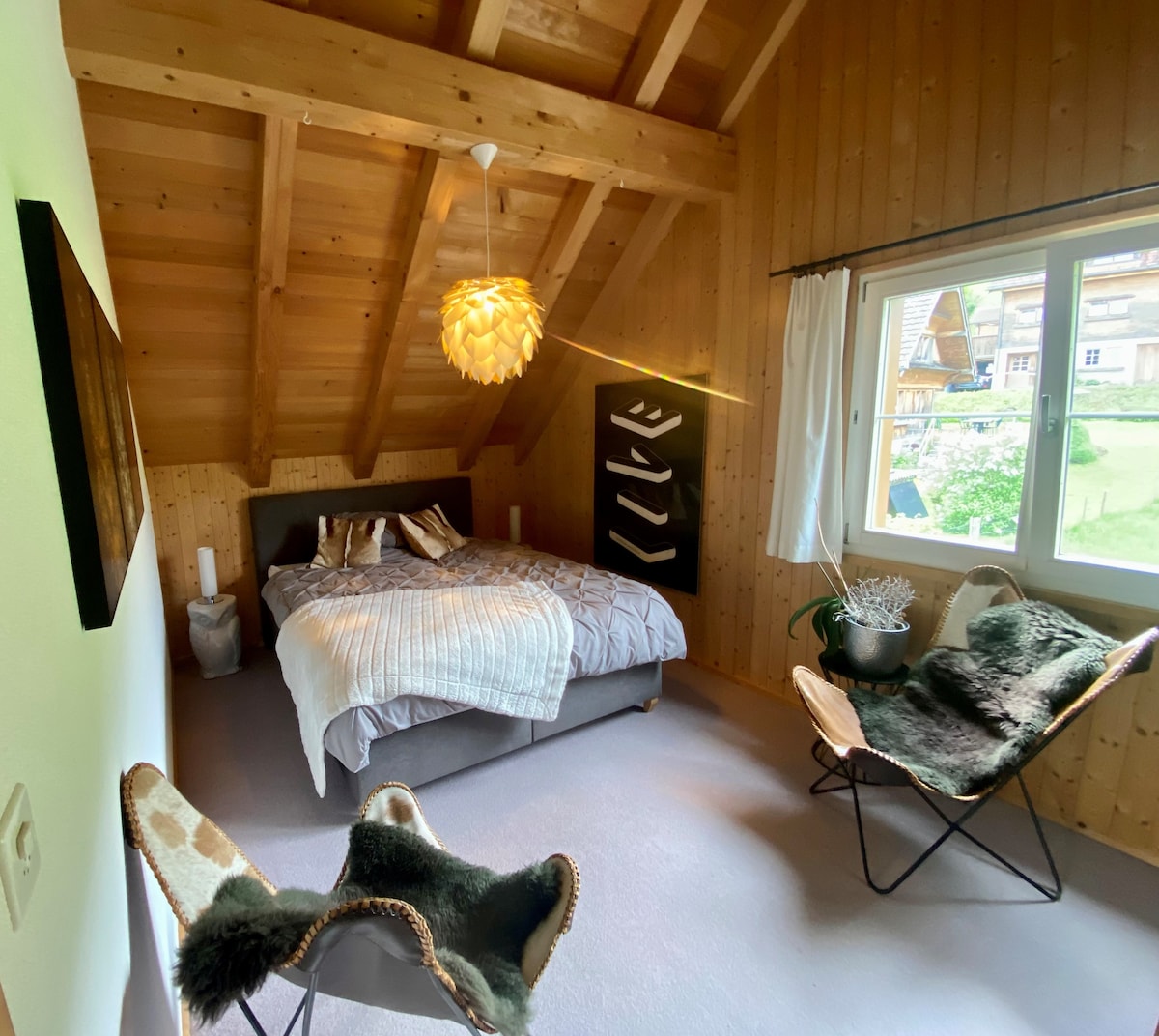 A guestroom features a spacious bed with neutral bedding and decorative pillows. Two distinctive chairs are placed near the window, allowing natural light to illuminate the wooden ceiling and walls. Soft textiles add comfort to the space, enhancing its inviting appearance.