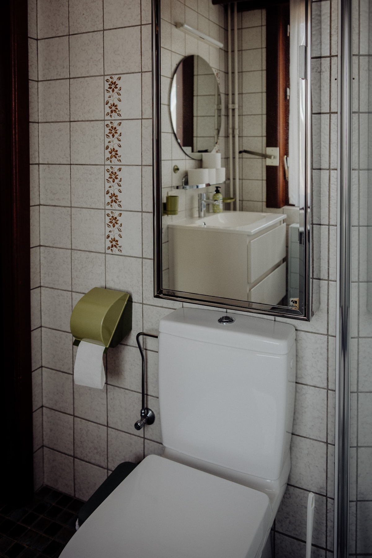 A clean bathroom space is shown, featuring a modern toilet and a rectangular mirror above a sleek vanity. The walls are tiled in light colors, with small decorative elements. A green toilet paper holder adds a subtle touch of color.