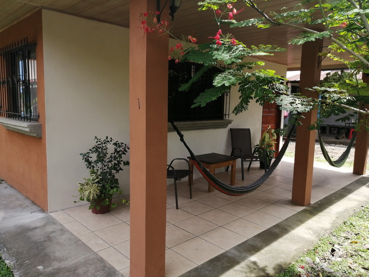 A shaded patio area is presented with tiled flooring, featuring a comfortable chair and a small table. A hammock is suspended between two posts, surrounded by greenery, including potted plants and vibrant trees, offering a relaxed outdoor space.