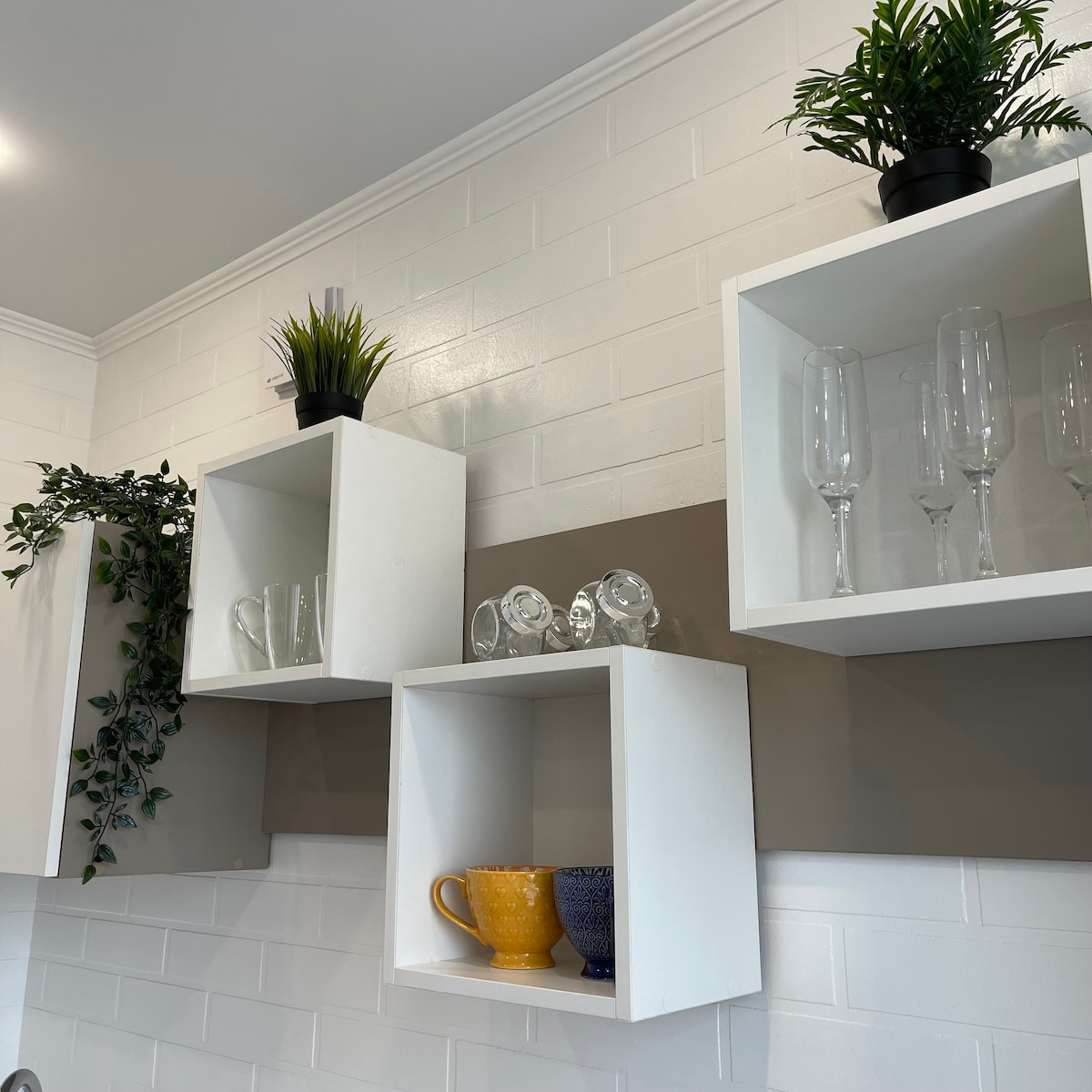Open shelving units are mounted on a white tiled wall, displaying a selection of glassware and decorative pots. Clear glass tumblers and champagne flutes are arranged alongside a yellow and blue ceramic cup, while lush greenery adds a touch of freshness.
