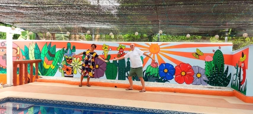 A vibrant mural adorns the wall beside a swimming pool, featuring colorful flowers, plants, and whimsical characters. The pool area is set against a backdrop of lush greenery, with a shaded seating area ensuring comfort while enjoying the outdoor space.