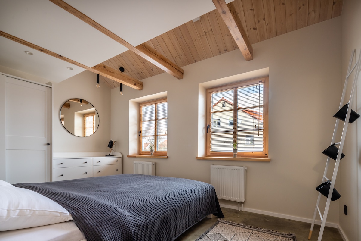 A bedroom featuring a spacious bed with a dark textured blanket. Natural light enters through two windows adorned with horizontal blinds. A round mirror is mounted above a dresser, and wooden beams accentuate the ceiling. A minimalist shelving unit is positioned against the wall.