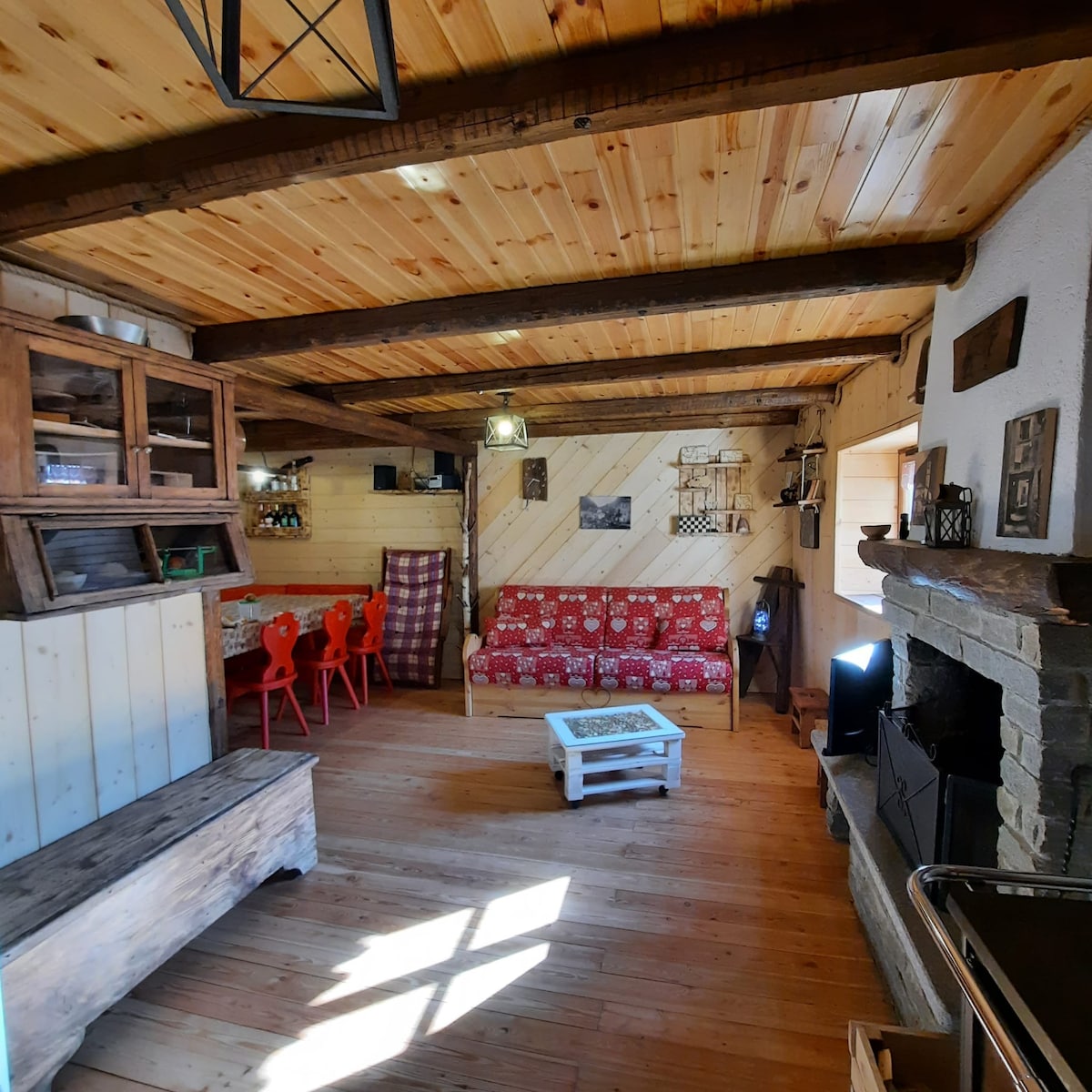 An inviting living area features a wooden ceiling and floor, complemented by natural light streaming through large windows. A cozy red patterned sofa is paired with a rustic wooden coffee table. A dining area with red chairs and a shelf displays decorative items, adding warmth to the space.