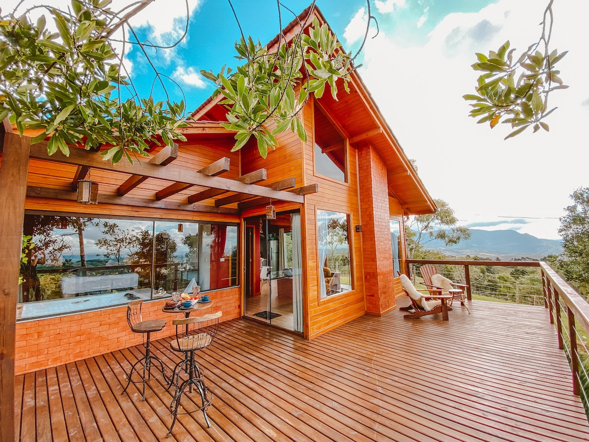 A wooden cabin features large glass walls and a spacious deck surrounded by trees. Outdoor furniture includes lounge chairs and a bistro table with chairs. The scenic view reveals rolling hills and blue skies, enhancing the peaceful ambiance of the space.