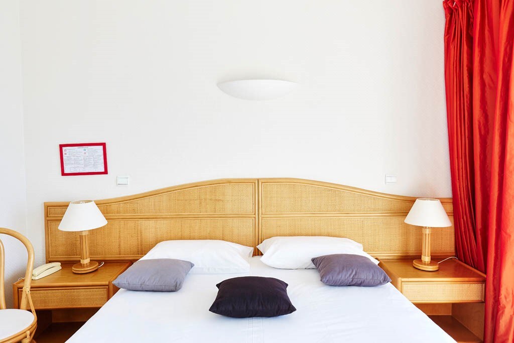 A light-filled sleeping area features a wooden headboard with bedside lamps on either side. The spacious bed is adorned with white linens and several accent pillows in varying shades. Bright red curtains frame the window, adding a pop of color to the serene environment.