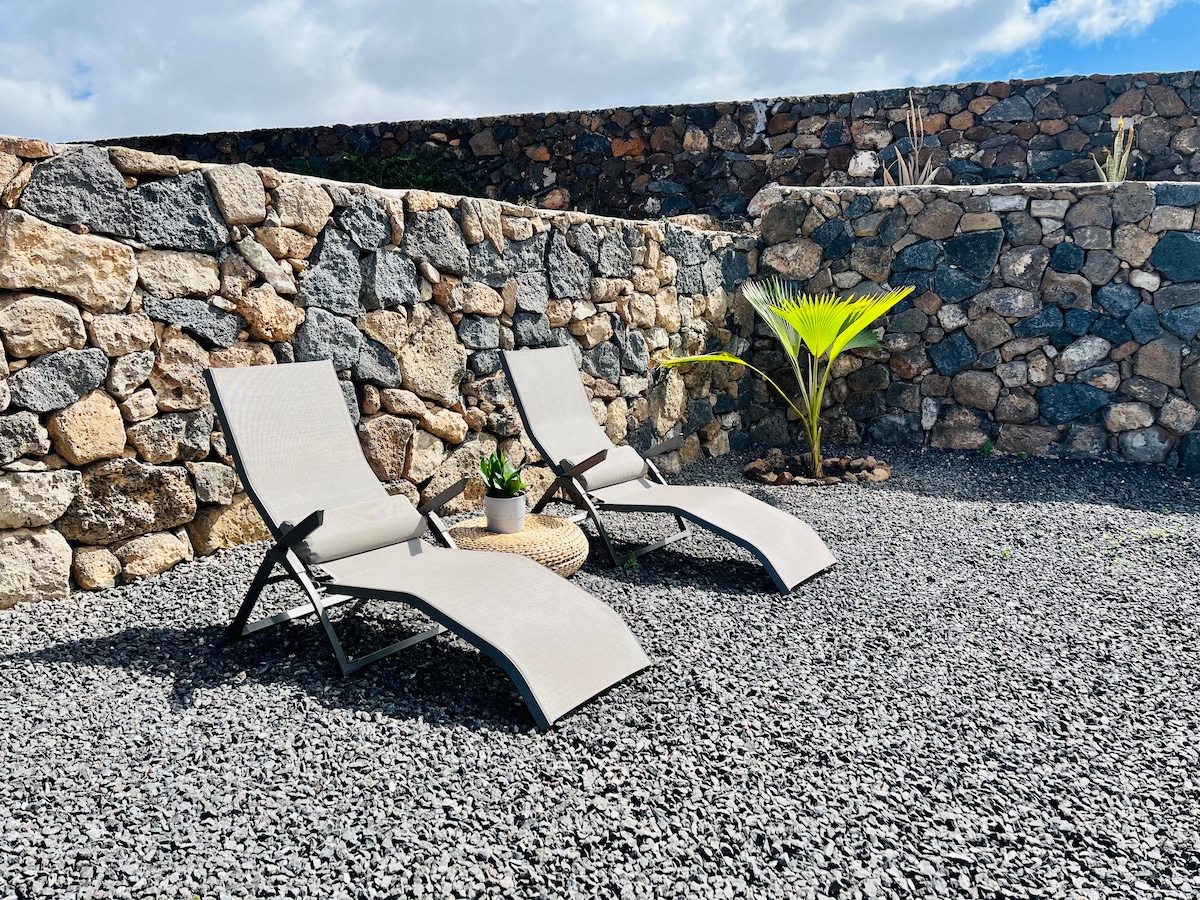 Two light grey lounge chairs are positioned on a gravel surface, accompanied by a small round table. A potted plant is placed on the table, and a stone wall surrounds the area, creating a private outdoor space. The sky is partly cloudy.