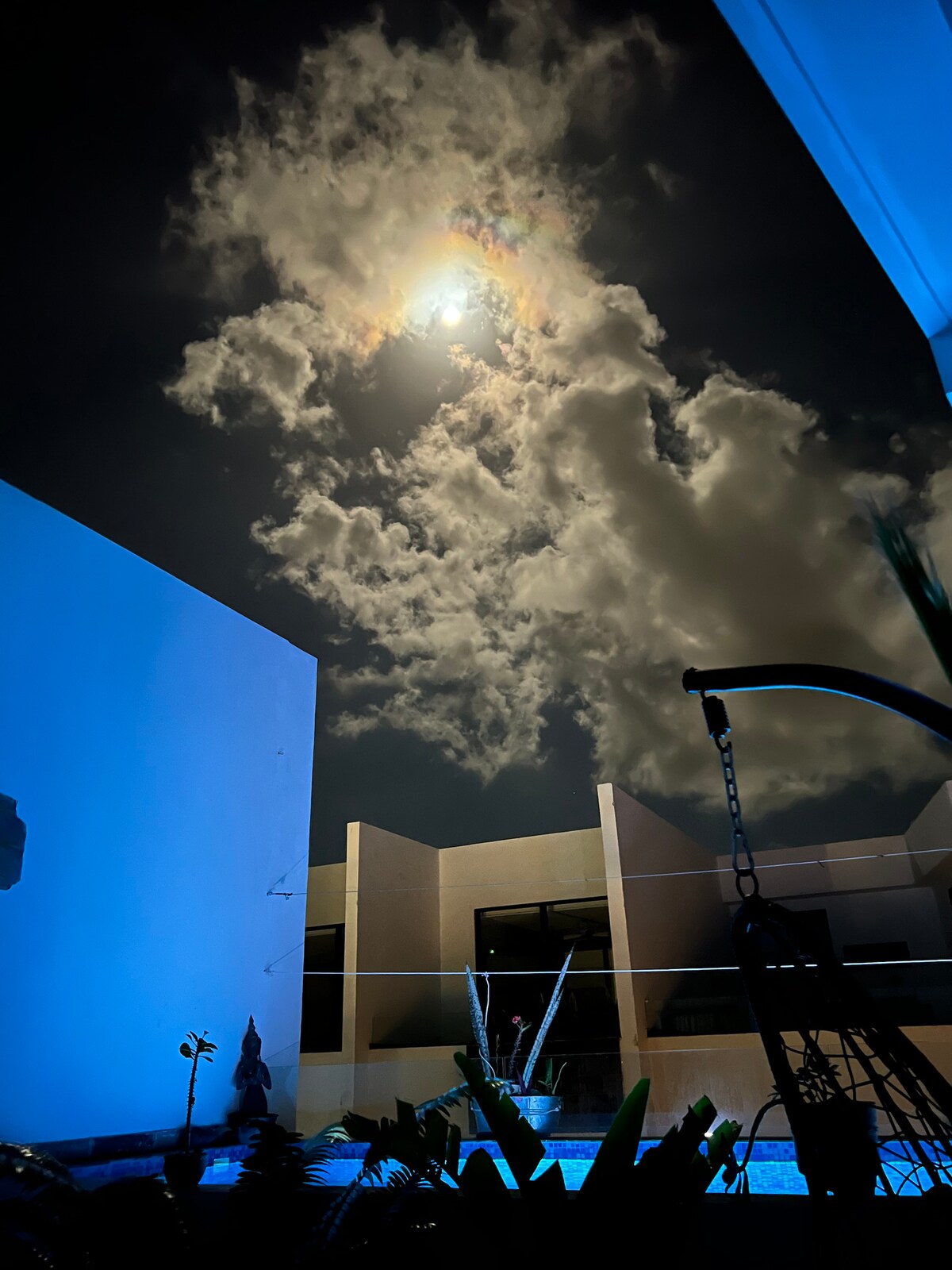 A tranquil outdoor area is illuminated by moonlight, highlighting a swimming pool surrounded by tropical plants. Soft clouds drift in the night sky above, while the smooth surfaces of modern architecture create a serene backdrop.