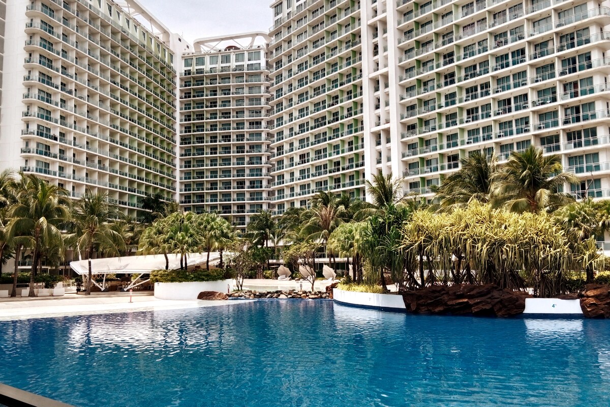 A large swimming pool is framed by palm trees and lush greenery, creating a serene outdoor space. Surrounding high-rise buildings are visible in the background, showcasing a mix of glass and white exteriors. The atmosphere appears calm and inviting.