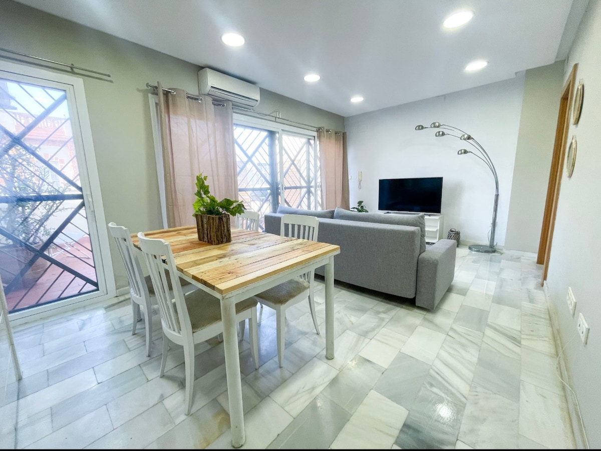 A modern living area features a wooden dining table surrounded by four chairs. A gray sofa and a floor lamp are positioned nearby. Natural light enters through large windows with sheer curtains, enhancing the space's brightness and warmth.