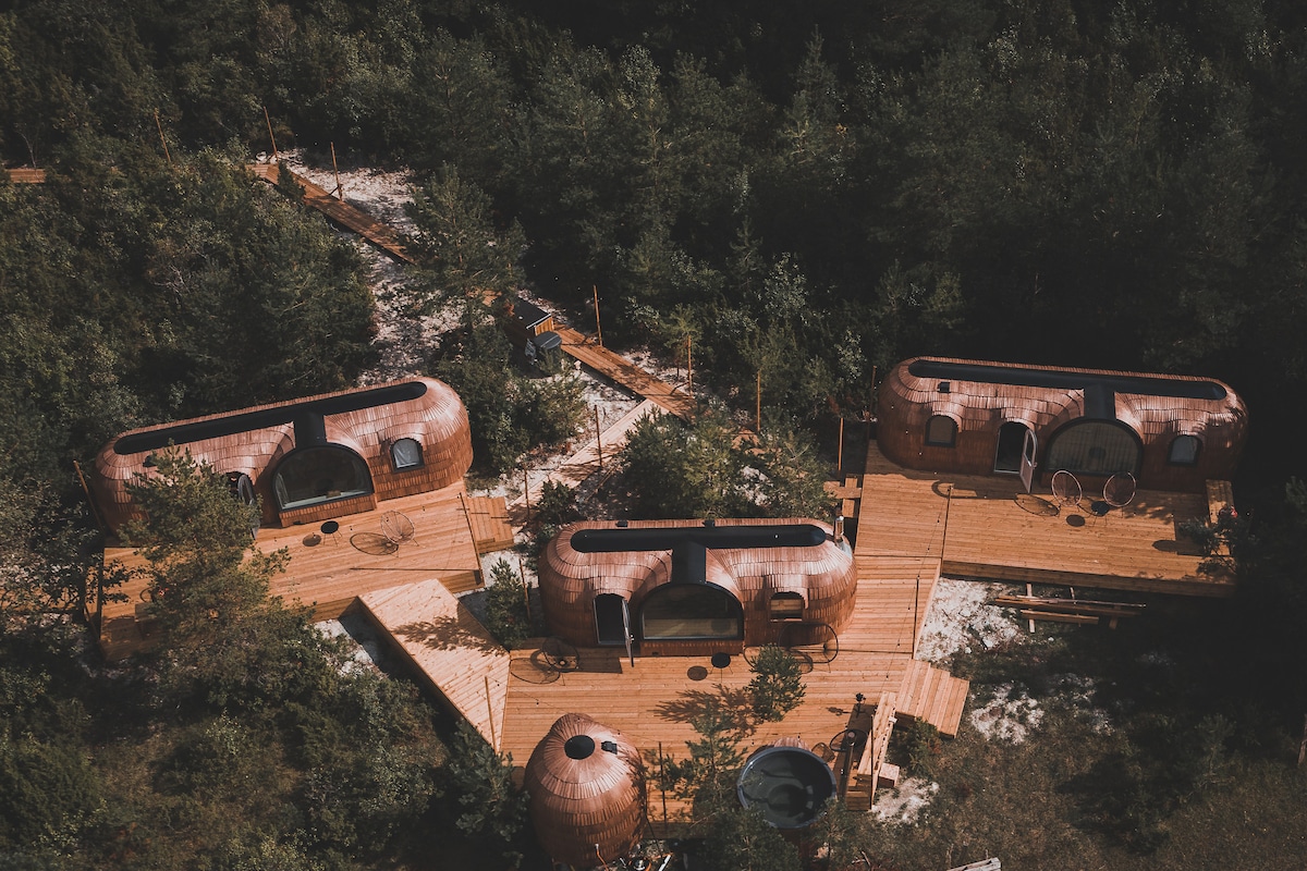An aerial view reveals multiple igloo huts nestled among lush greenery, interconnected by wooden pathways. Each hut features curved copper exteriors, large windows, and outdoor terraces. The serene surroundings highlight the retreat's natural setting, complemented by a hot tub visible in the foreground.