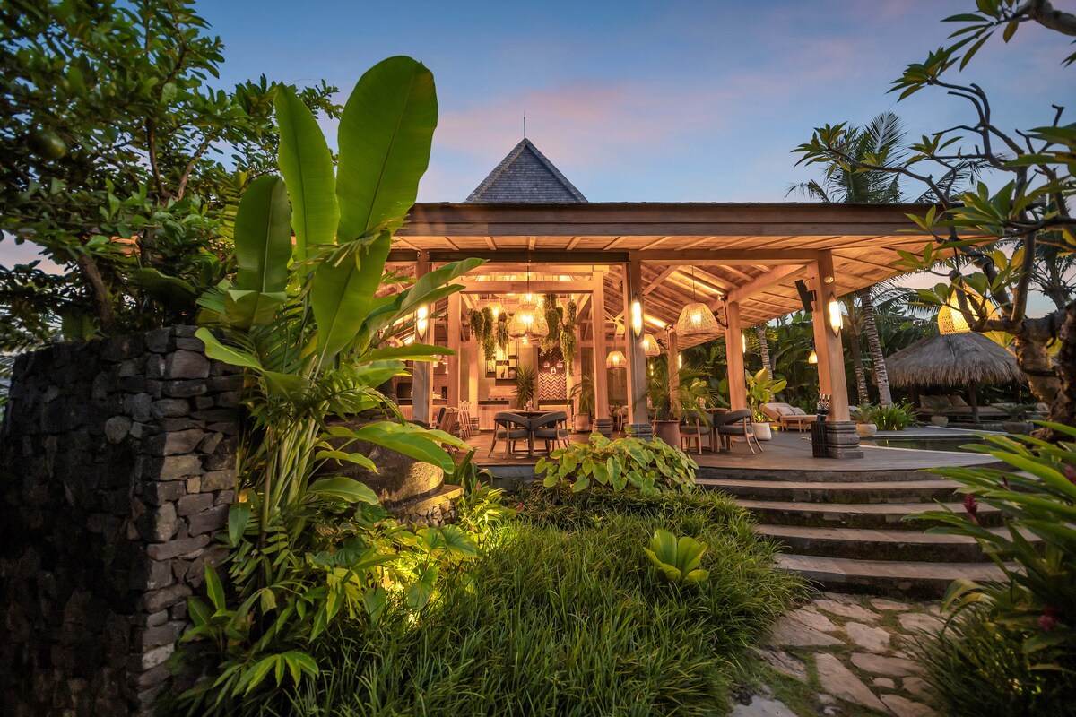 The exterior of a spacious villa is framed by lush greenery, featuring a wooden structure with inviting outdoor seating. Soft lighting accentuates the entrance, while stone pathways lead through the vibrant garden, creating an atmosphere that blends nature with comfortable living.