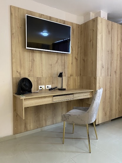 A compact workspace is integrated into the wooden wall, featuring a wall-mounted television above a simple desk. A reading lamp rests on the desk next to electrical outlets. A neutral-colored chair with metal legs complements the modern design.