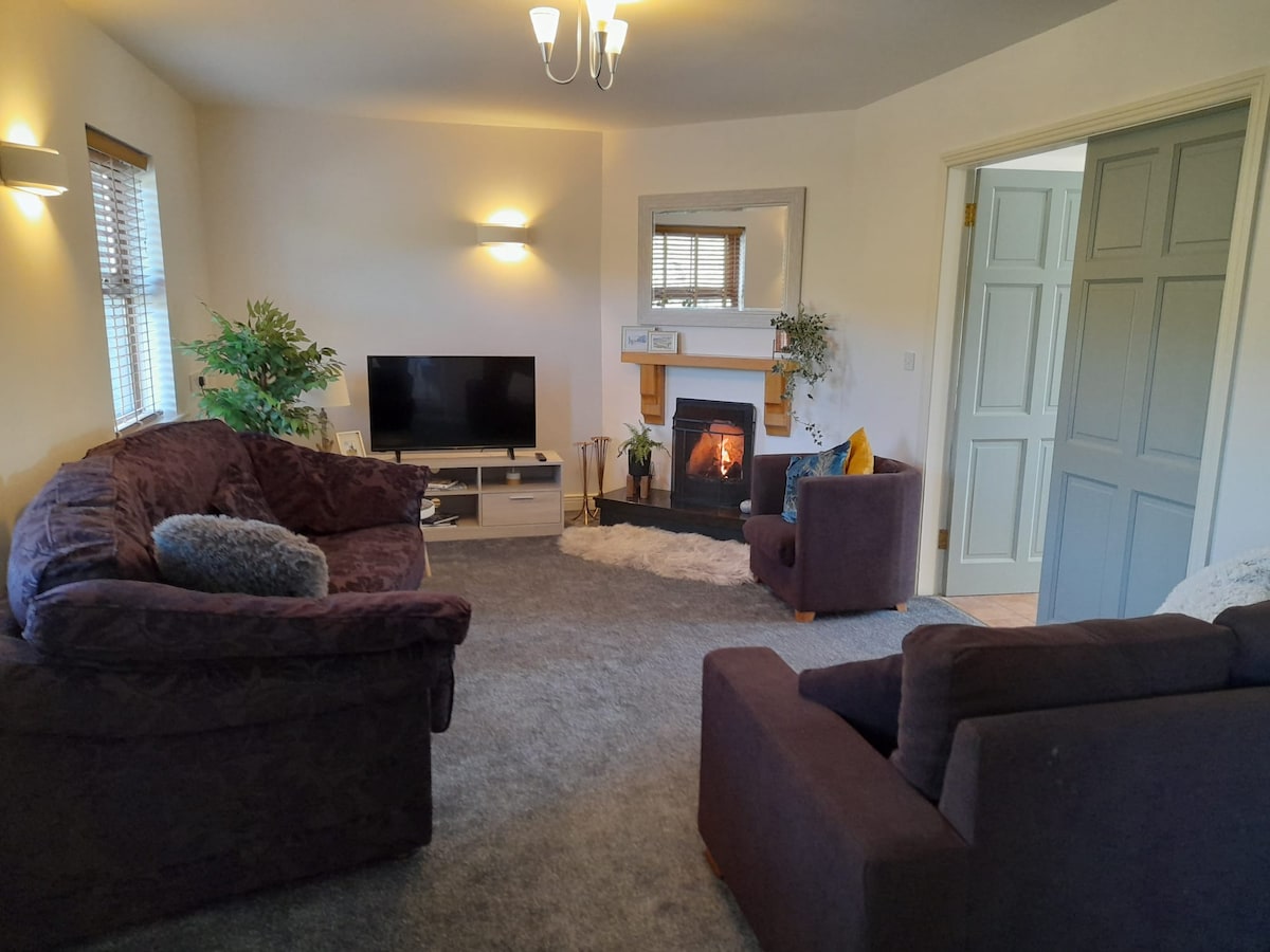 A cozy living room is presented featuring two comfortable sofas and a small armchair. A television is positioned against the wall, while a fireplace adds warmth to the space. Natural light enters through the windows, enhancing the room's welcoming atmosphere.