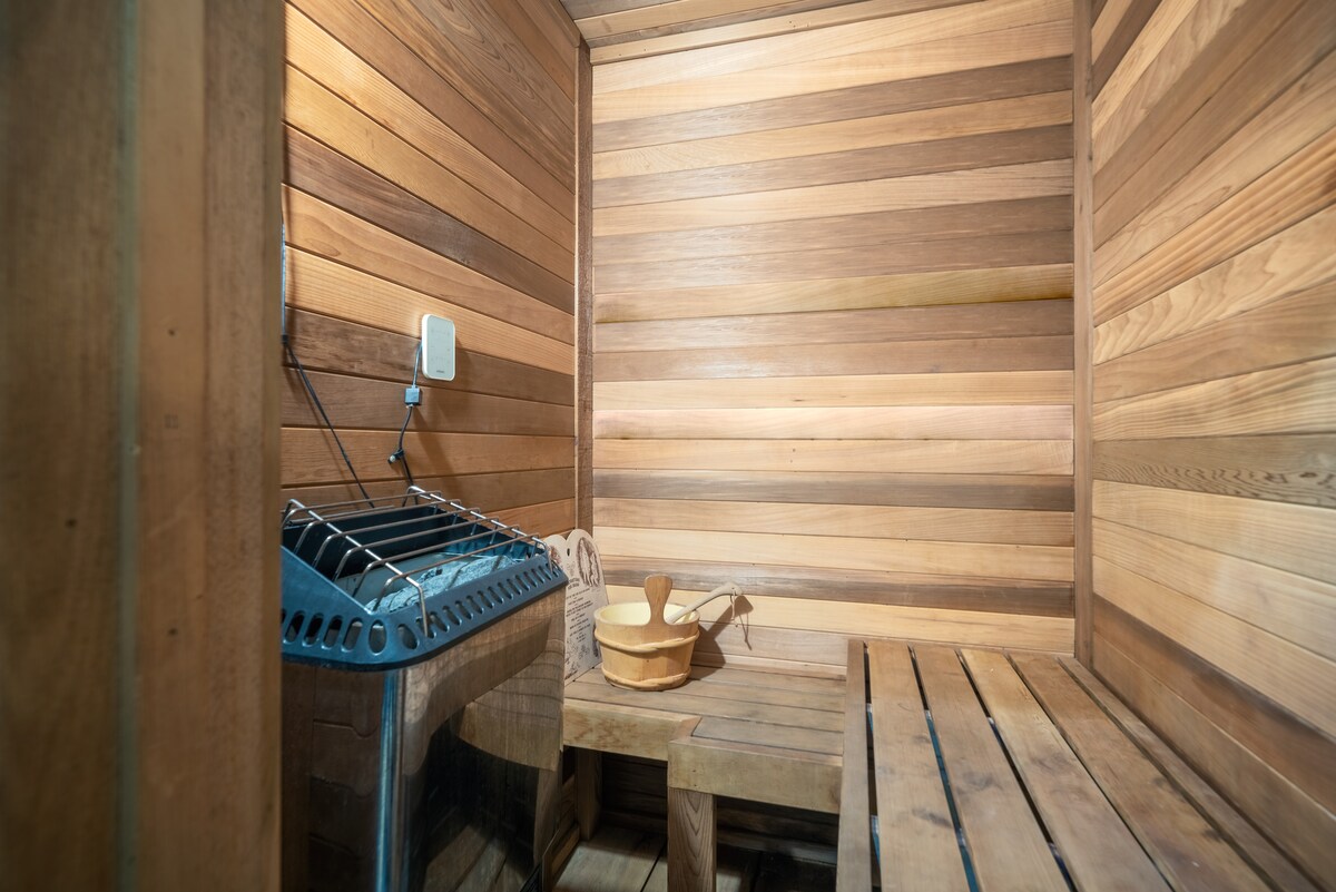 The sauna features natural wood paneling, creating a warm atmosphere. A metal heater is visible alongside a wooden bench and a small table holding a bucket and ladle, offering a space for relaxation and wellness.