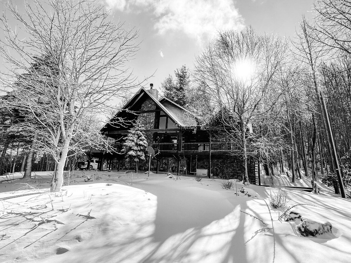 A rustic mountain cabin is surrounded by a snowy landscape, showcasing a mix of stone and timber construction. Sunlight breaks through the trees, casting shadows on the untouched snow in the yard. The cabin features multiple levels and a wide balcony overlooking the serene winter environment.