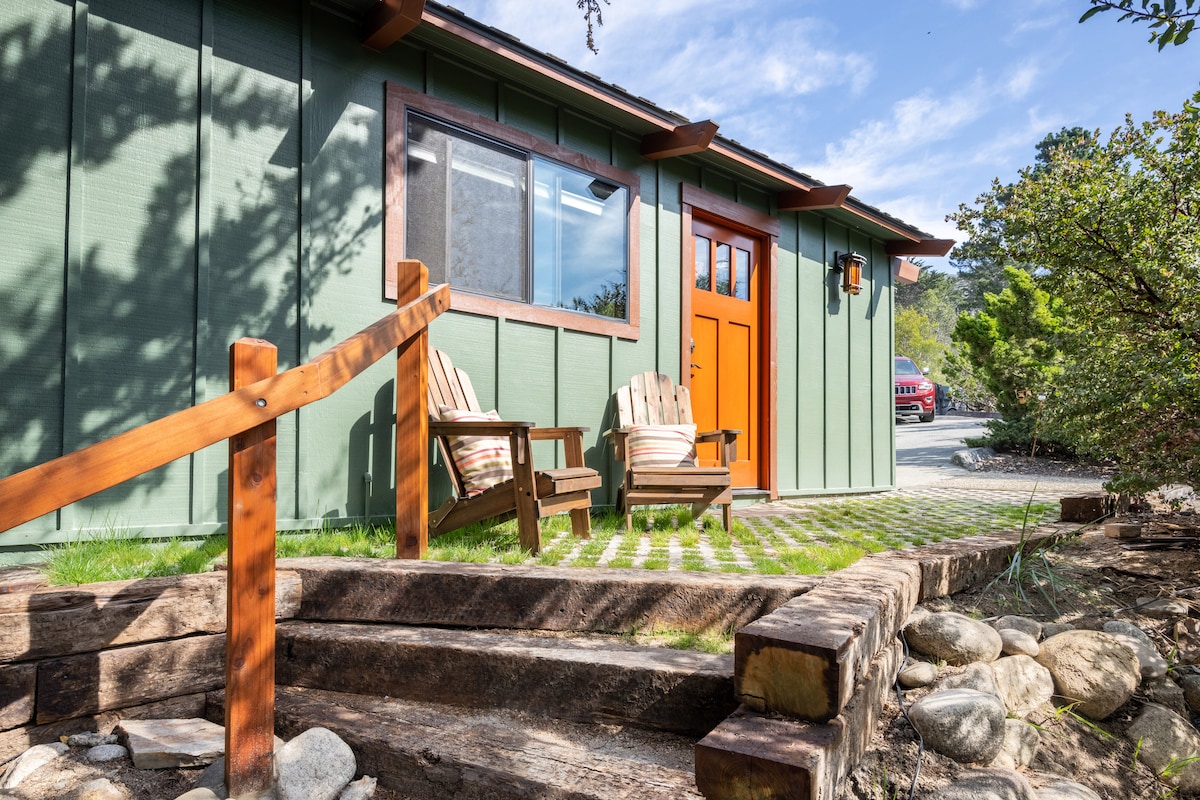 The exterior of a rustic bungalow features a welcoming red door flanked by two wooden chairs, each with striped cushions. A stone pathway leads to the entrance, surrounded by landscaped greenery and boulders, providing a serene outdoor setting.