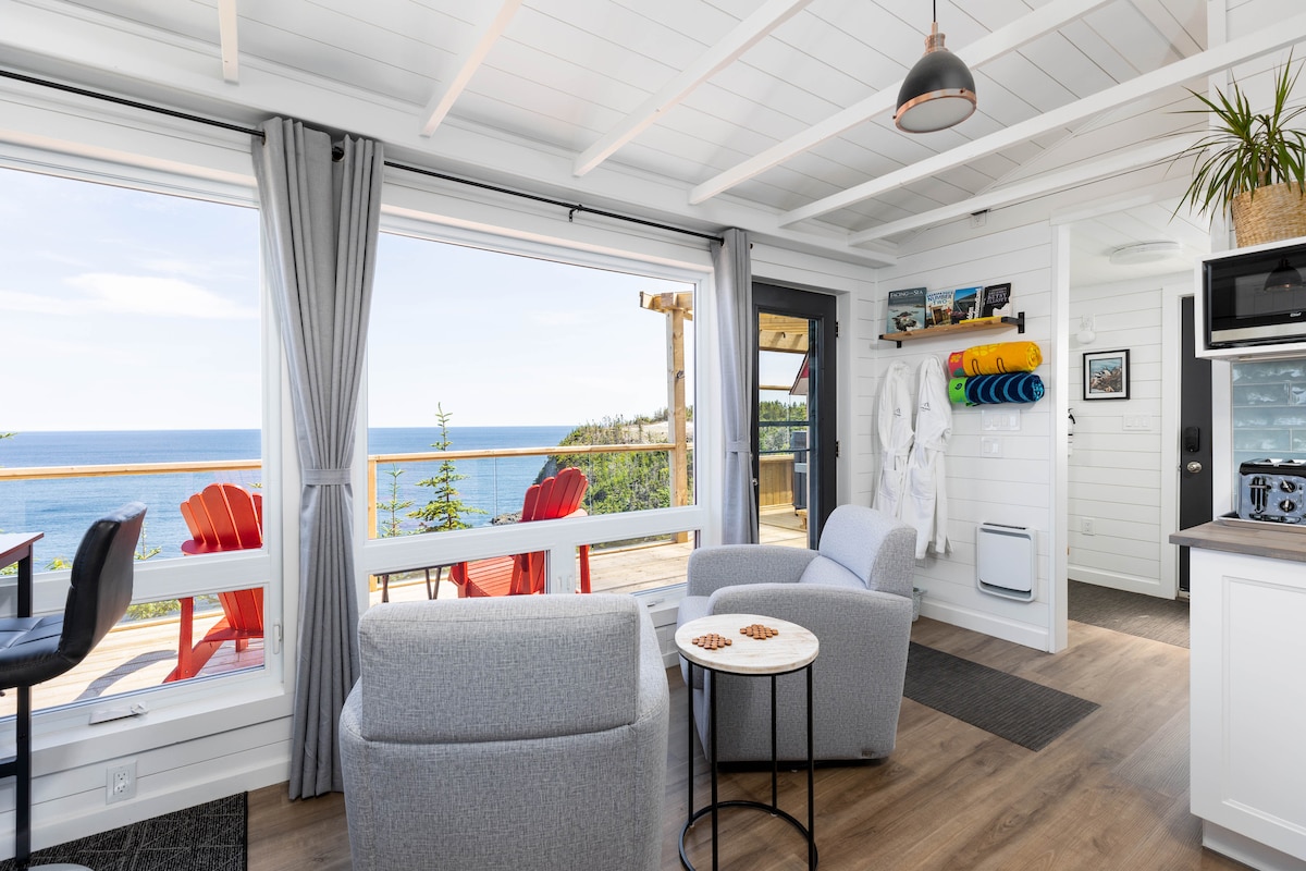 A bright seating area features two armchairs and a small side table, with beach-inspired decor. Large windows frame the ocean view, allowing natural light to fill the space. Coastal colors are evident in the outdoor seating area visible on the deck.