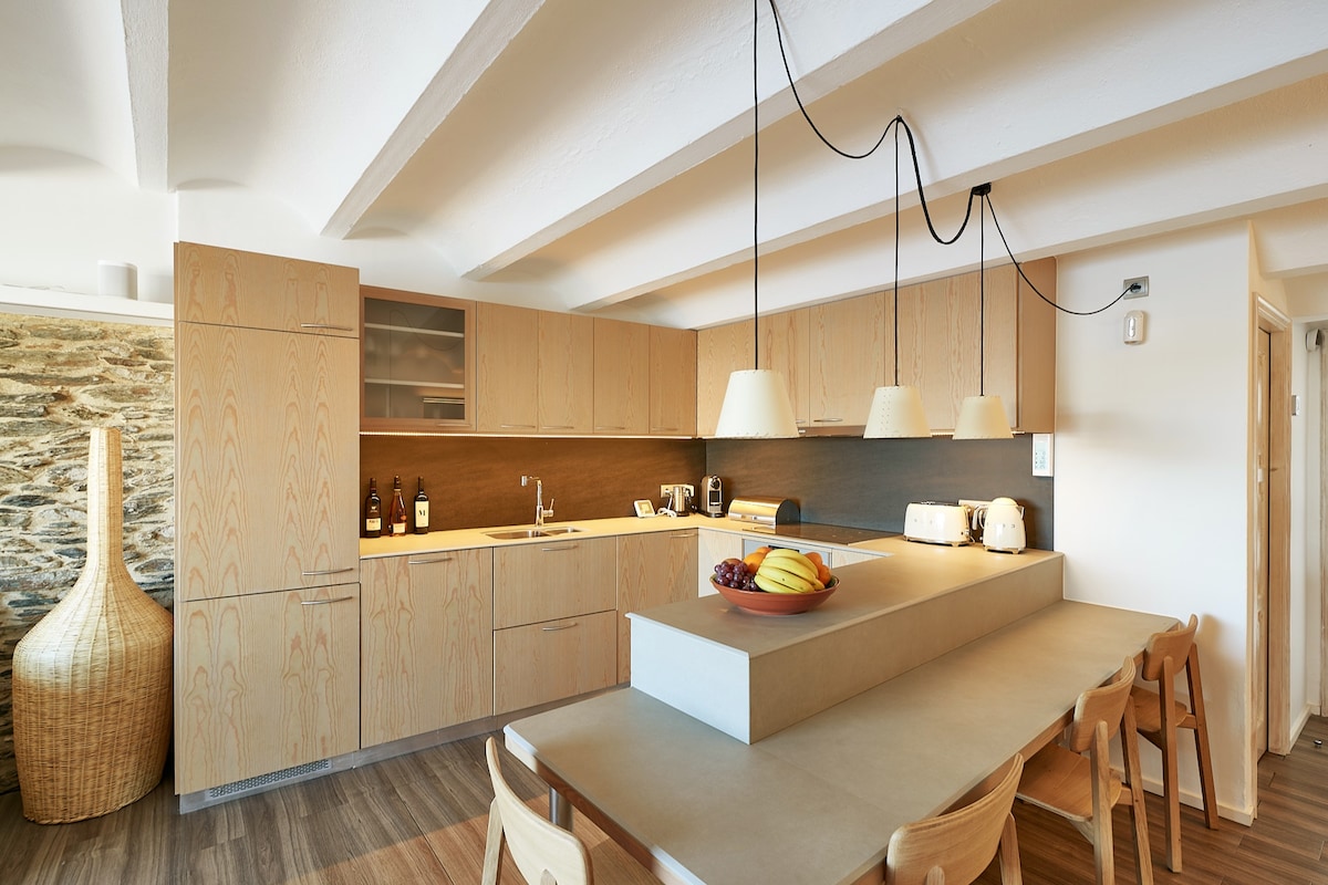 The modern kitchen features wooden cabinetry and a central island with a fruit bowl. Overhead pendant lights illuminate the space, enhancing the sleek design. A spacious dining table with wooden chairs is positioned beside the island, creating an inviting area for meals and gatherings.