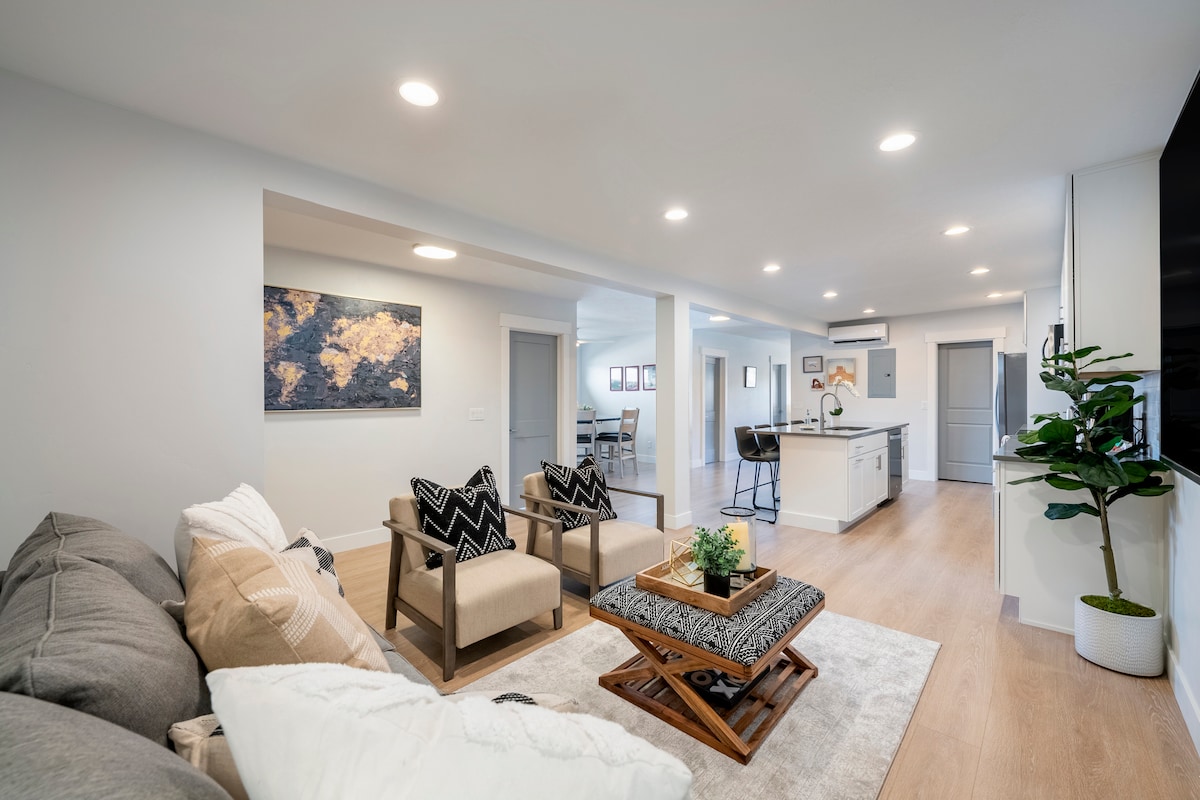 A spacious living area features a comfortable gray sofa and two armchairs, accented with patterned cushions. A coffee table with greenery is positioned on a soft area rug. The open layout connects to a bright kitchen, illuminated by recessed lighting and natural light from nearby windows.