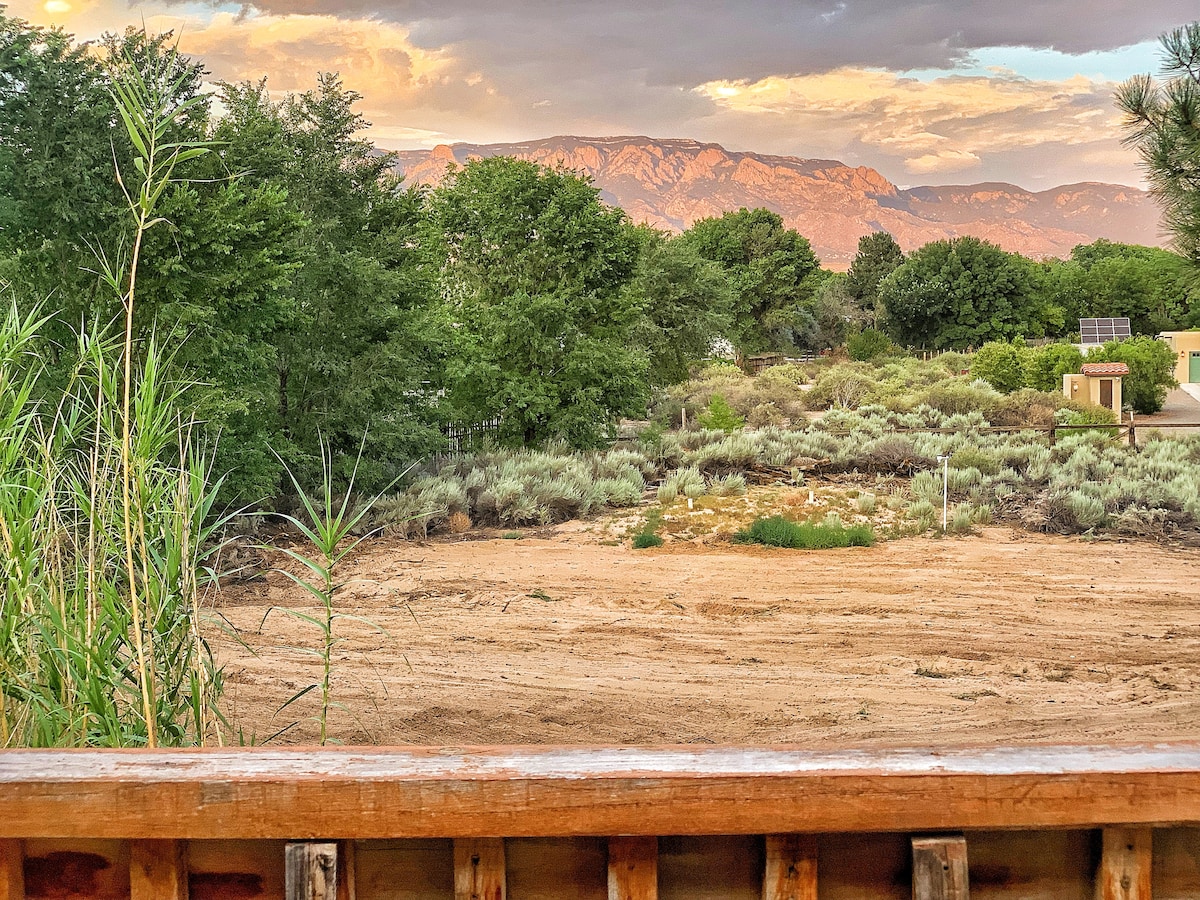 A natural landscape unfolds beyond the wooden railing, featuring shrubs and earthy tones under a soft evening sky. The Sandia Mountains rise in the background, framed by greenery. The scene captures the serene atmosphere of the surrounding area, inviting moments of reflection and tranquility.