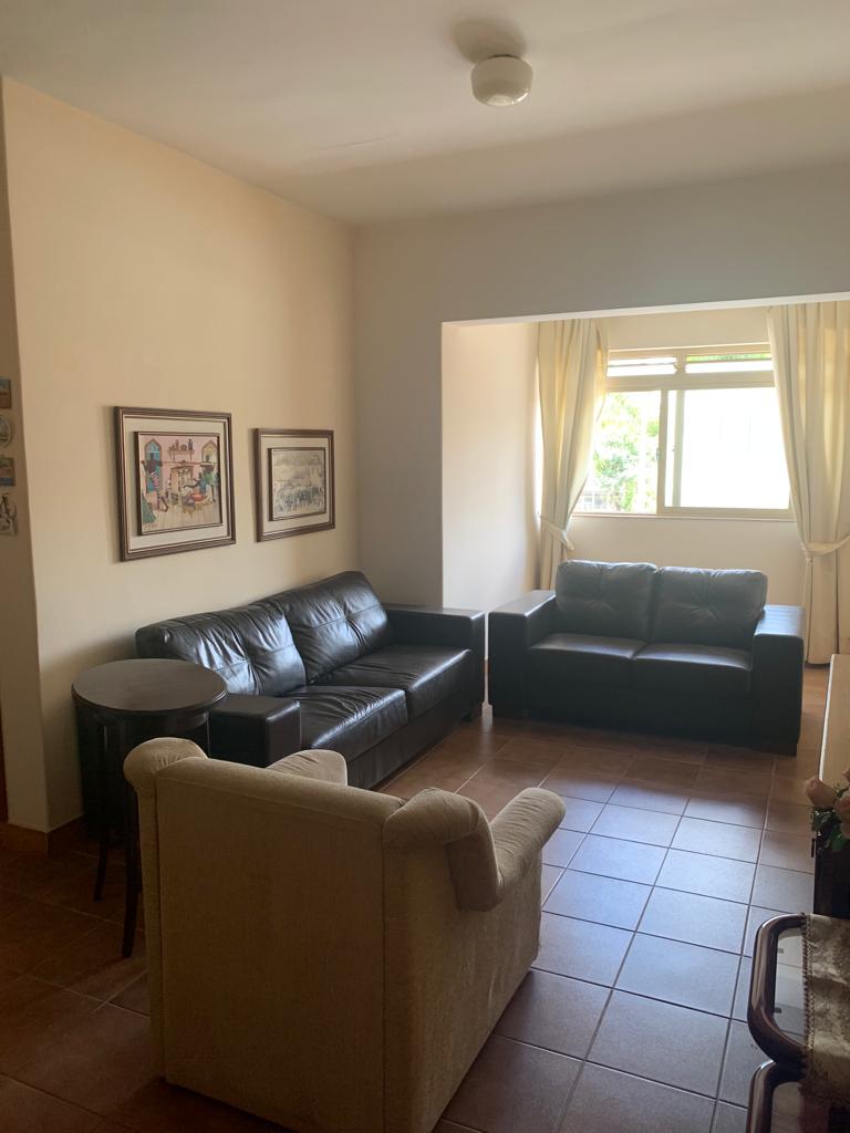 A living area features a dark leather sofa and a matching armchair, arranged to promote conversation. Natural light enters through a large window, which is adorned with soft curtains. Two framed art pieces hang on the wall, adding subtle character to the space.
