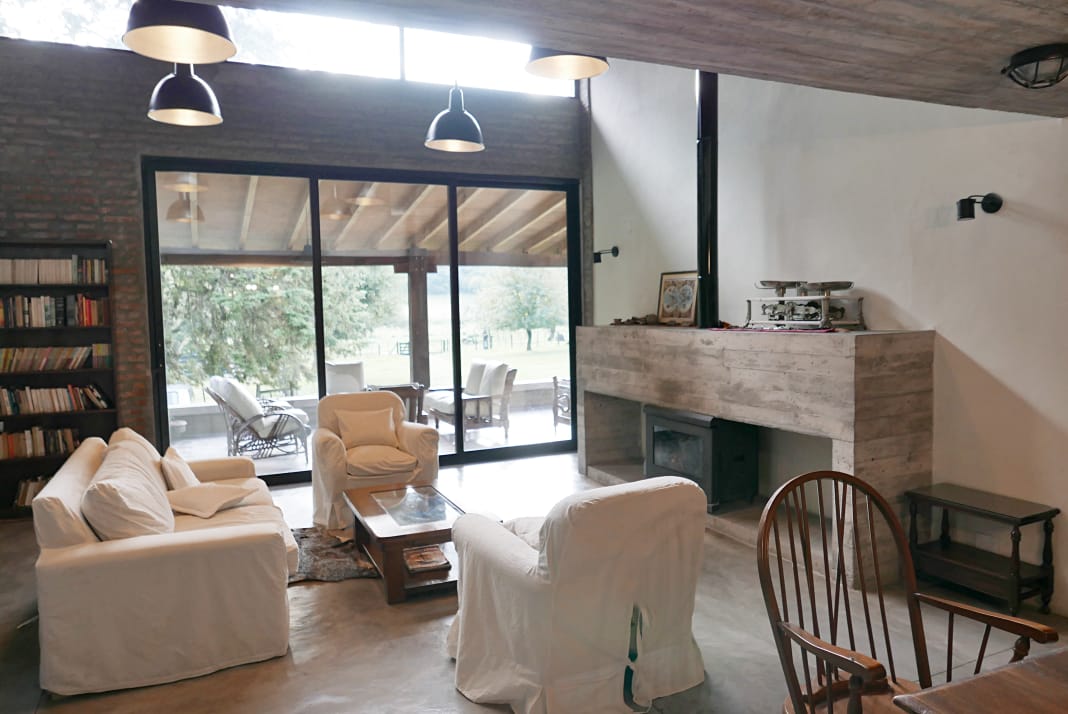 A spacious living area features white upholstered seating arranged around a wooden coffee table. Large glass doors open to a view of the outdoors, while a contemporary fireplace is located against a textured wall. A bookshelf is positioned along one side.