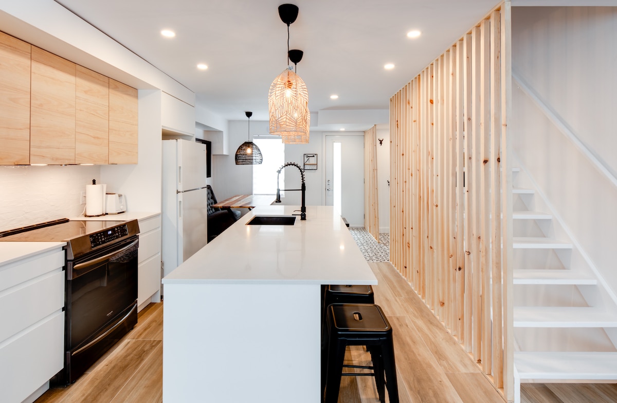 An open-concept kitchen and dining area features a central island with seating and modern appliances. Pendant lighting hangs above the island, while wooden accents create an inviting space. Natural light fills the room through large windows, complemented by light-colored cabinetry and a cozy color scheme.