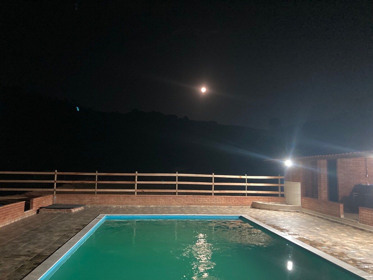 A serene outdoor area features a swimming pool illuminated by soft lighting. The night sky is visible above, showcasing a full moon casting a gentle glow over the surroundings. A wooden fence borders the space, enhancing the tranquil atmosphere.