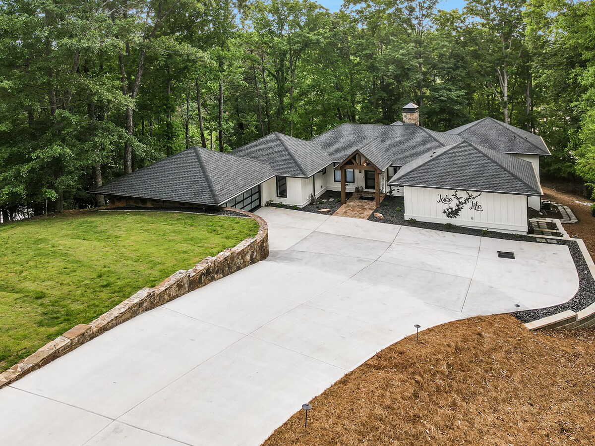 The exterior of a contemporary single-story home is showcased, featuring a combination of stone and siding. A spacious driveway leads to a welcoming entrance. Surrounding greenery provides a natural backdrop, enhancing the peaceful and private setting of the residence.