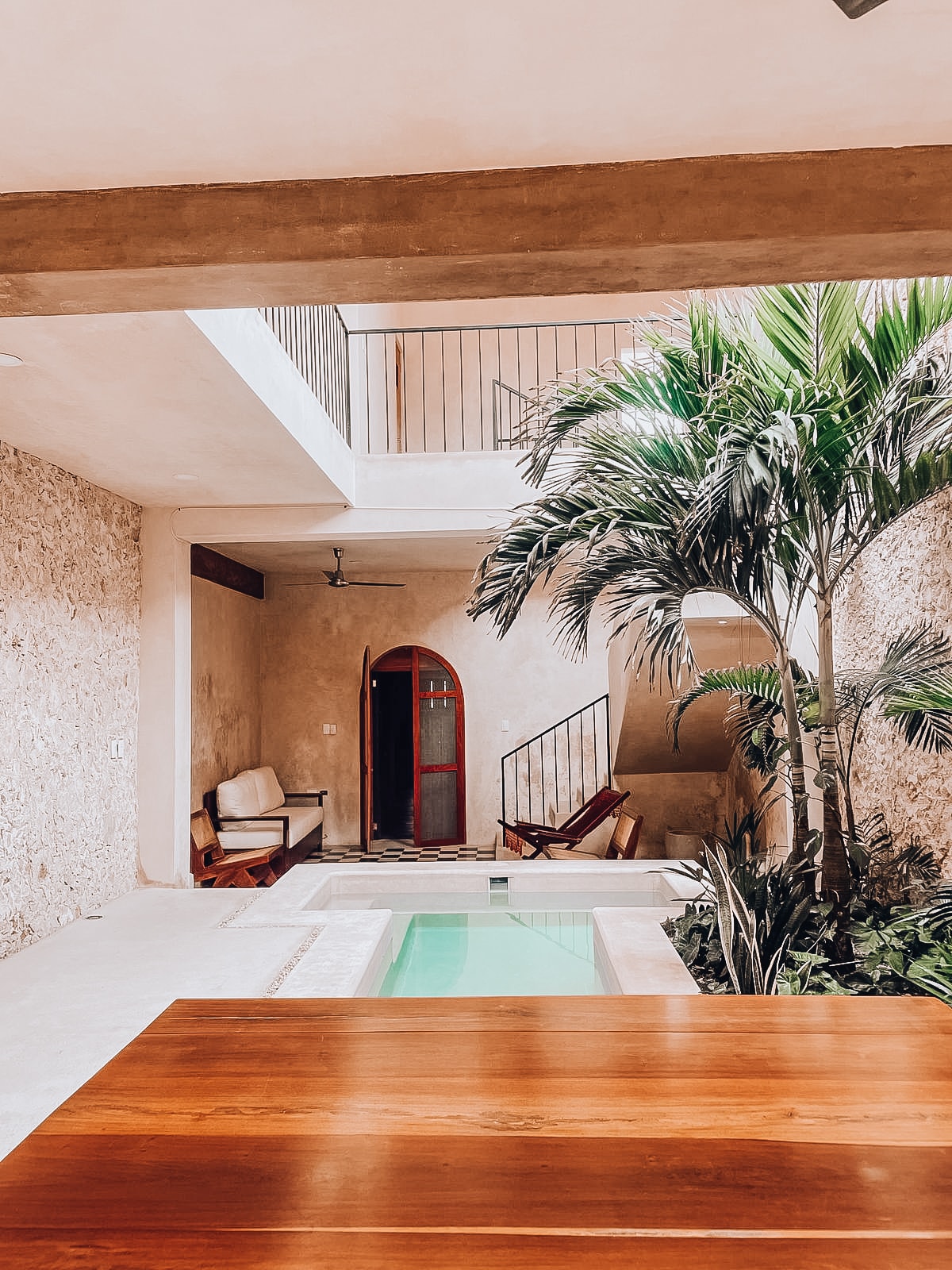 A serene courtyard is highlighted by a small pool surrounded by lush greenery. Wooden lounge chairs are positioned nearby, and natural textures of stone and plaster create a calm ambiance. An arched doorway leads to the area, enhancing the open and inviting design.