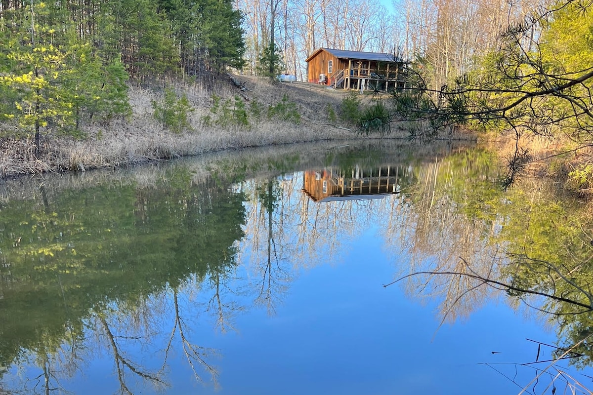 A tranquil pond reflects the wooden cabin in the background, surrounded by lush greenery and trees. The calm water captures the peaceful setting of the property, emphasizing the serene atmosphere of the little bear cabin.