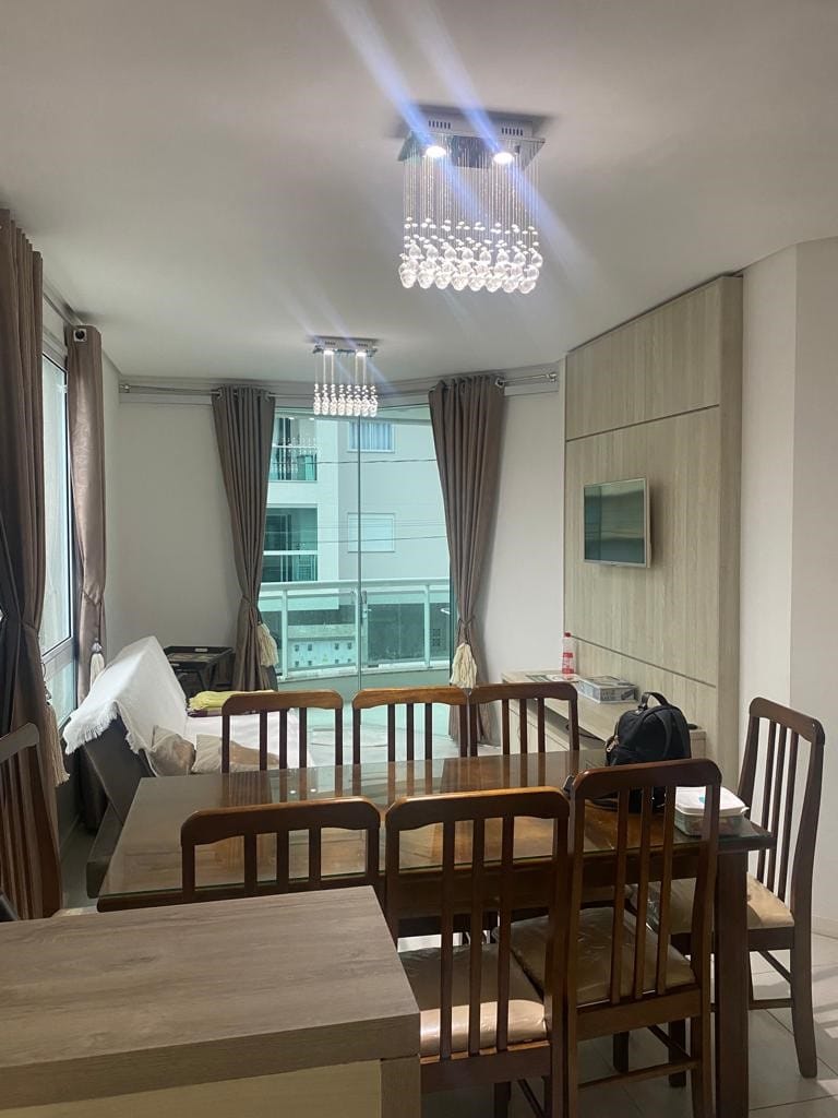 A dining area features a wooden table surrounded by several chairs, with a view provided by large windows that allow natural light to fill the space. Modern light fixtures hang from the ceiling, and soft curtains frame the windows, enhancing the room's warmth.