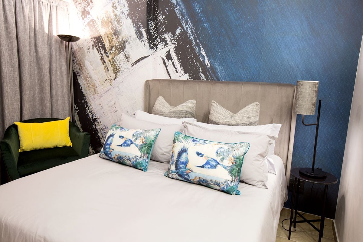 A well-furnished queen room features a spacious bed dressed with white linens and decorative pillows showcasing aquatic designs. A modern lamp stands beside the bed, while a green accent chair adds a pop of color against a textured blue wall.