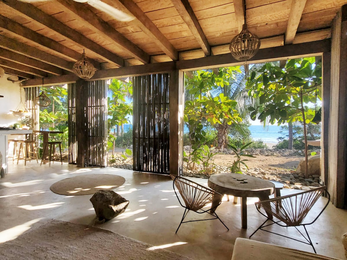 The open main room features a blend of natural light and greenery, with views of the ocean visible through wooden slatted doors. A round natural fiber rug is placed on the floor, accompanied by a low circular wooden table and two inviting seating chairs.