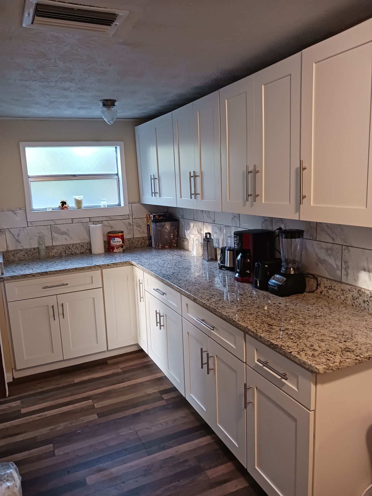 The kitchen is equipped with white cabinetry and a speckled granite countertop. Small appliances including a coffee maker and blender are present. A window provides natural light, highlighting the modern design and functional layout of the space.