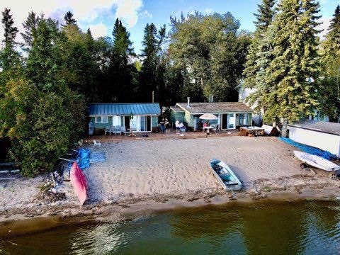 Two beachfront cottages are positioned side by side on a sandy shore, surrounded by lush trees. The cottages feature outdoor seating and are complemented by a canoe and a small boat moored nearby. The serene lake reflects the natural setting.