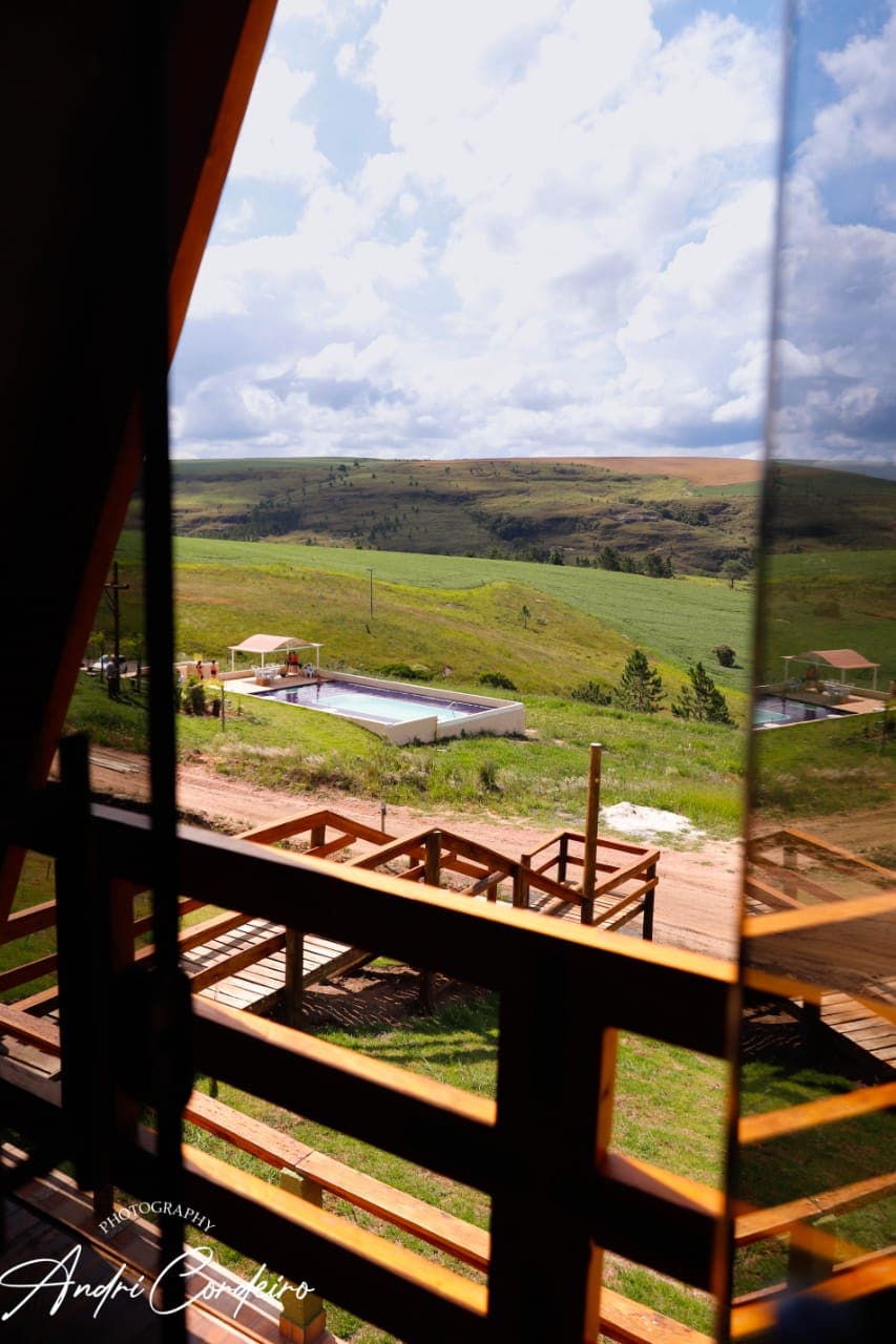 A scenic view is captured from the balcony, showcasing lush green hills and open fields under a partly cloudy sky. A swimming pool is visible below, surrounded by natural landscapes, while wooden decks connect outdoor spaces.