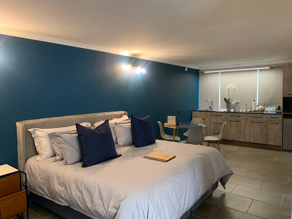 A spacious sleeping area features a king bed with multiple decorative cushions against a back wall painted in a deep blue. A cozy seating area includes a small table and two chairs. A fully equipped kitchenette is visible in the background, with natural light streaming through a window.
