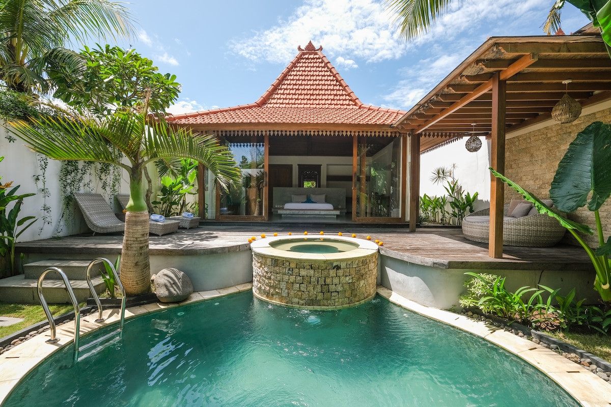 A private pool is framed by lush greenery, featuring a circular stone seating area. The villa’s traditional roof design is visible in the background, complemented by a spacious wooden deck. Comfortable lounge chairs are positioned near the pool, creating a serene outdoor space.