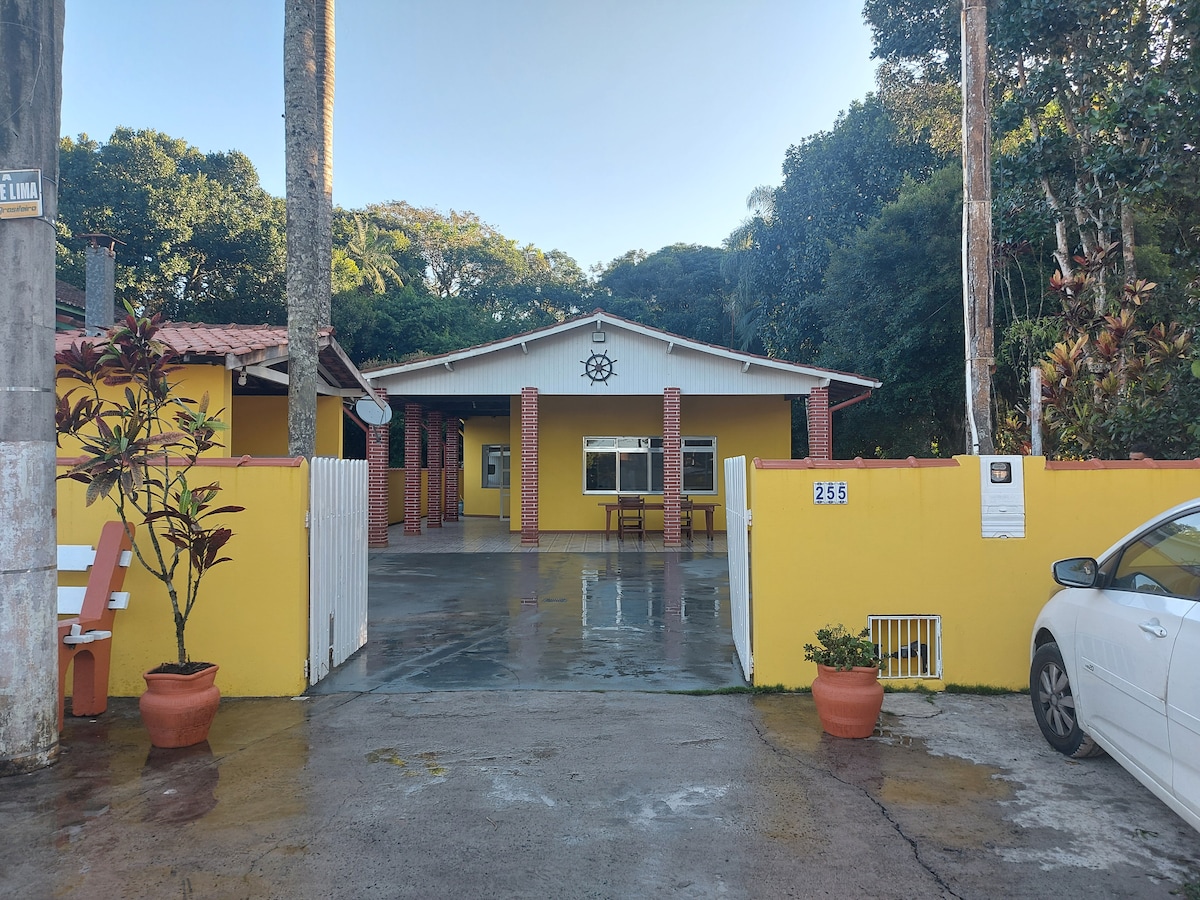 The exterior features a bright yellow façade with red brick accents. A spacious driveway is visible, surrounded by potted plants. A covered porch area is presented, with a wooden table and chairs under the home's overhang. Lush greenery frames the property.