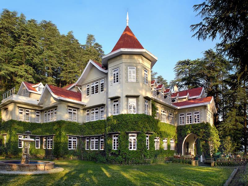 A historical building surrounded by lush greenery showcases intricate architecture with multiple gabled roofs and numerous windows. The façade is adorned with climbing plants, and a decorative fountain is visible in the foreground, adding to the serene outdoor environment.