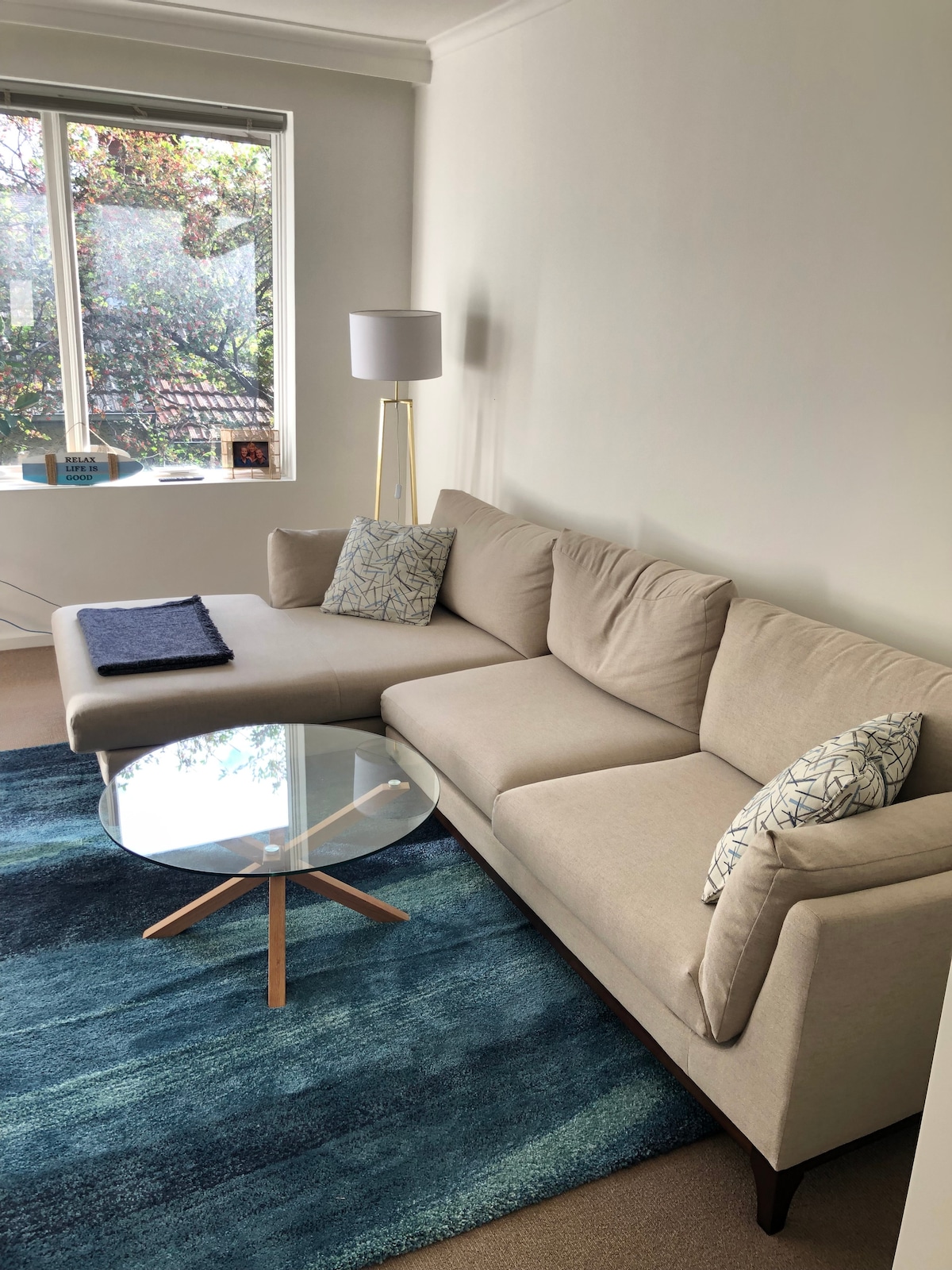 A light-filled living area features a beige sectional sofa adorned with patterned cushions. A glass coffee table with wooden legs sits on a textured blue rug. Natural light streams in from a window, creating an inviting space for relaxation.