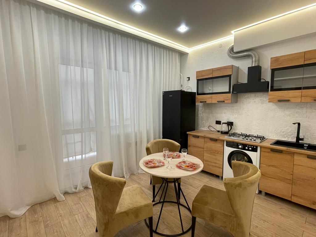 A modern kitchen area features light wood cabinetry and sleek black accents. A round dining table with four chairs is set with plates and glasses. Large white curtains allow soft light to filter in through the window, highlighting the open space.