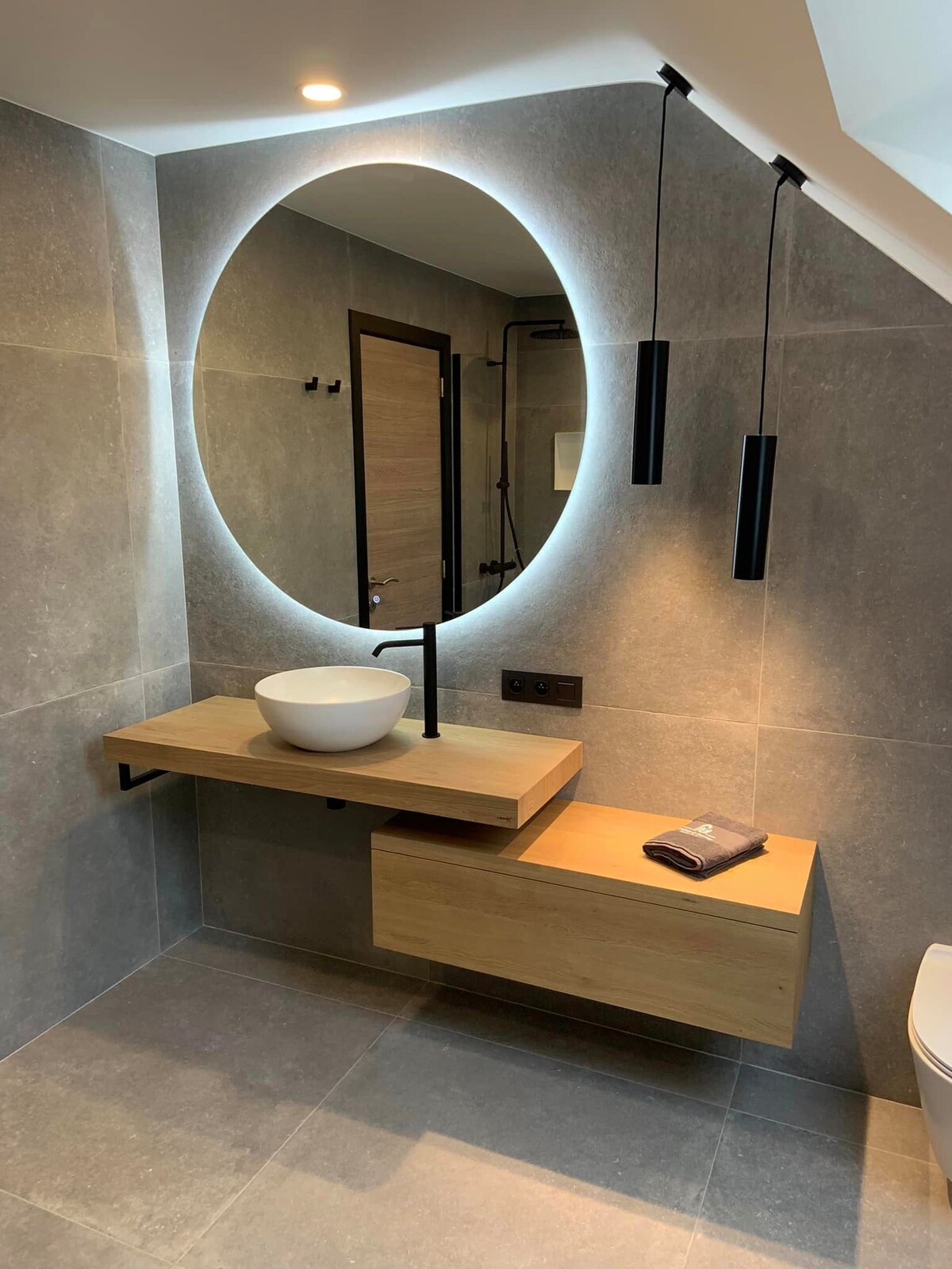 A modern bathroom features a circular mirror with backlighting, enhancing the sleek design. A wooden vanity holds a bowl sink, and a drawer is integrated below. Minimalist black fixtures and pendant lights contribute to the clean aesthetic, while the floor is tiled in a neutral grey.